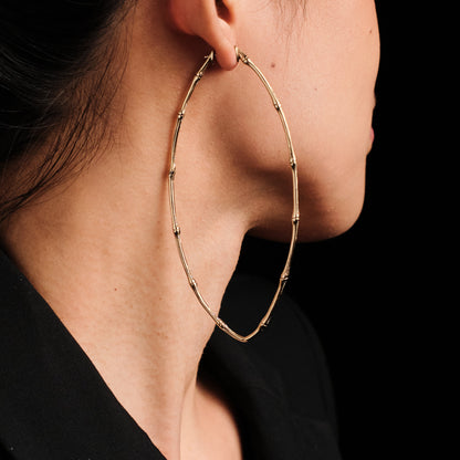 A close-up side profile of a woman wearing a large gold hoop earring with a textured, beaded bamboo-like design. The background is solid black.