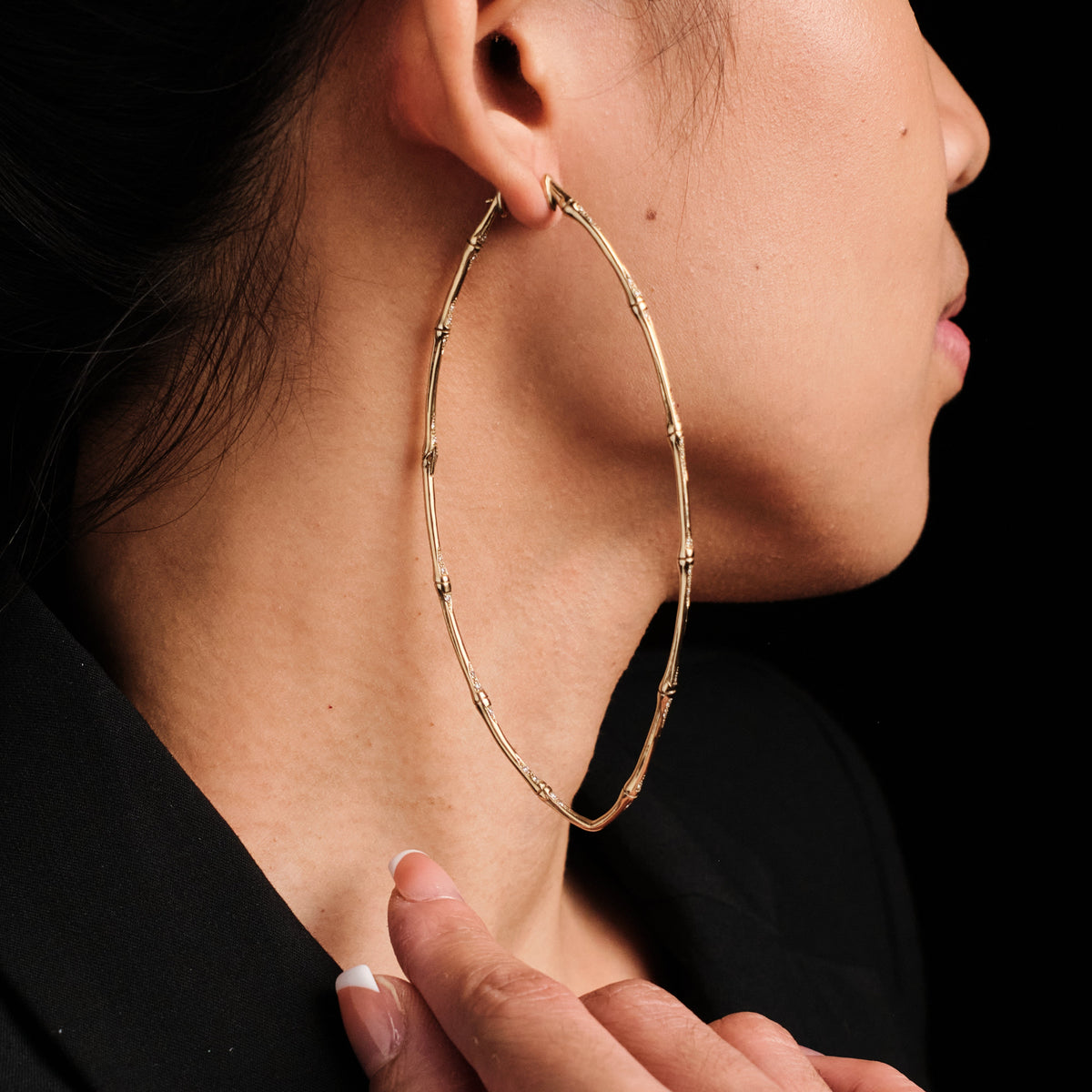 A close-up side profile of a woman wearing a large gold hoop earring with a textured, segmented bamboo-like design. She is wearing a black top against a solid black background.